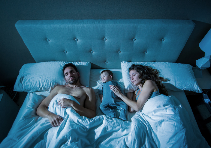 A baby between his mom and dad on the bed with pillows and covers all around him.
